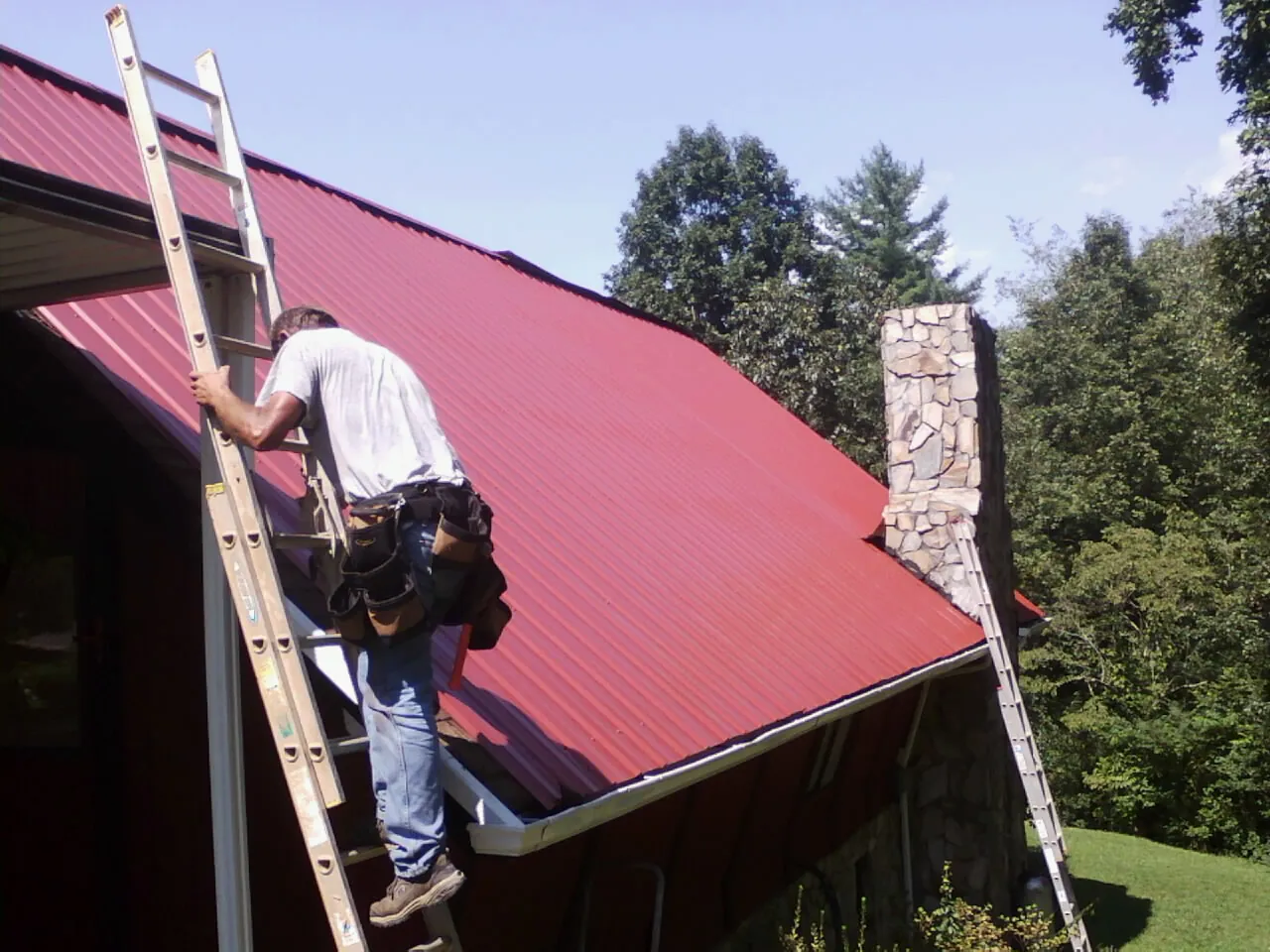 Roofer on a ladder beside a red metal roof and stone chimney — Soffit & Fascia Repair by Roofing Brothers in Servia